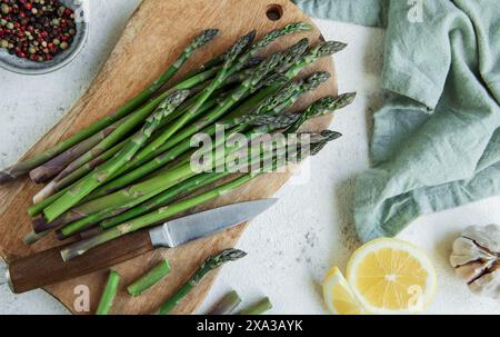 Gli asparagi verdi freschi giacciono su un tagliere di legno accanto a un coltello da cucina, con fette di limone e spicchi d'aglio nelle vicinanze. Il tagliere è acceso Foto Stock