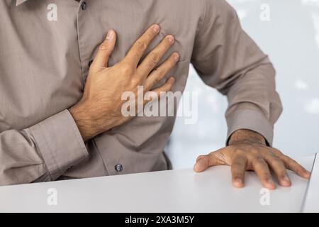 Primo piano di un uomo che soffre di dolore toracico, che tiene il petto con una mano e riposa l'altra su un tavolo. Foto Stock