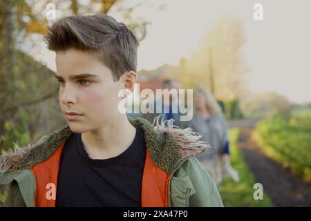 Ragazzo adolescente a piedi nella strada di campagna, i genitori in background Foto Stock