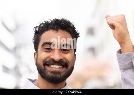 Un uomo sorridente con i capelli ricci sta alzando il pugno in un gesto di successo o celebrazione su uno sfondo urbano sfocato. Foto Stock