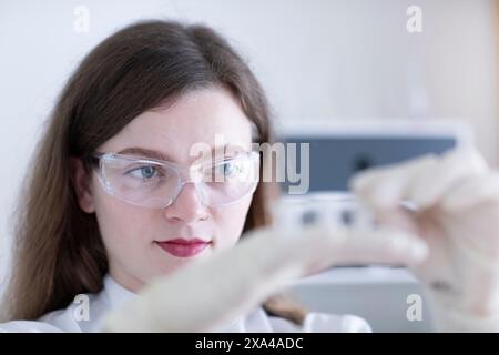 Una donna concentrata che indossa occhiali di sicurezza sta esaminando una fiala in laboratorio. Foto Stock