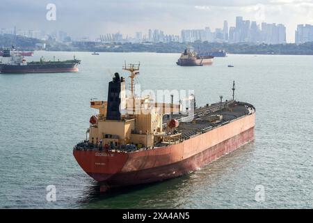 Una grande nave da carico rossa e nera è ancorata in un porto affollato con molte altre navi nelle vicinanze e uno skyline della città sullo sfondo sotto un cielo nuvoloso. Foto Stock