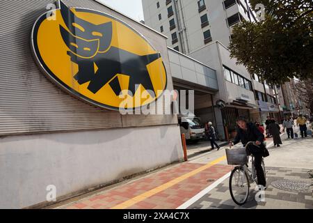 Una donna corre in bicicletta davanti a un grande logo della Yamato Delivery Company in un deposito a Ginza, Tokyo, Giappone. Foto Stock