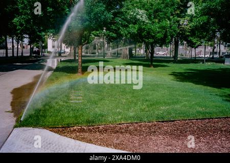 Un irrigatore innaffia un lussureggiante parco verde, creando un arcobaleno visibile nella nebbia sullo sfondo di alberi e un cielo limpido. Foto Stock