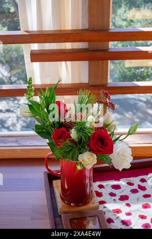 Un vivace bouquet di fiori con rose rosse e tulipani bianchi in una tazza rossa, collocata su una superficie di legno vicino ad una finestra con tende a vista e una vista del verde esterno. Foto Stock