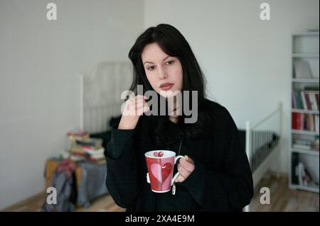 Una donna con i capelli scuri regge una tazza rossa, in piedi in una stanza con una libreria bianca sullo sfondo. Foto Stock