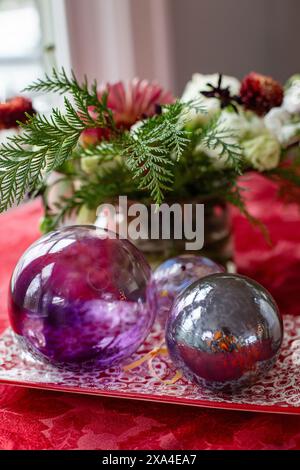 Un ambiente da tavolo vivace e festoso caratterizzato da due sfere decorative riflettenti che poggiano su un portamonete in pizzo scarlatto, accompagnato da una lussureggiante composizione floreale con fogliame verde. Foto Stock