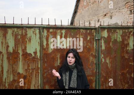 Una donna si trova di fronte a un cancello di metallo arrugginito con punte sulla parte superiore, con un muro di mattoni sullo sfondo, che crea una sorprendente espressione facciale. Foto Stock