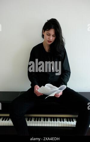 Una giovane donna siede su una panchina per pianoforte, vestita con un abbigliamento nero, sfoggia spartiti musicali con un'espressione premurosa. Foto Stock