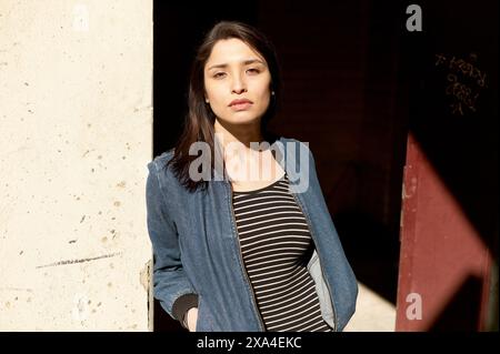 Una donna è illuminata da luce naturale, appoggiata a una parete e indossa una giacca in denim su un top a righe. Foto Stock