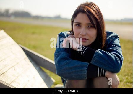 Una donna con capelli castani di media lunghezza siede all'aperto, poggia il mento sulle braccia incrociate, con uno sfondo morbido con erba e un cielo soleggiato. Foto Stock