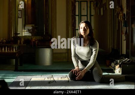 Una donna è seduta sul pavimento all'interno di una stanza con la luce del sole che filtra attraverso una finestra, gettando ombre intorno a lei; sembra contemplativa. Foto Stock