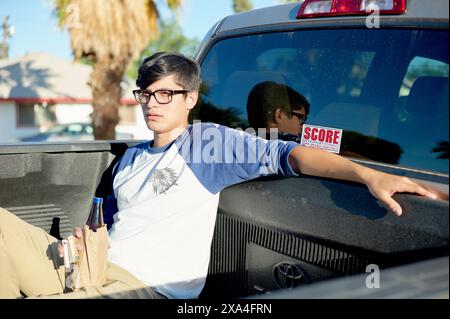 Un giovane con gli occhiali che indossa una camicia blu e grigia e che tiene in mano una bottiglia blu si siede comodamente sul retro di un pick-up, con una strada residenziale e palme sullo sfondo. Foto Stock