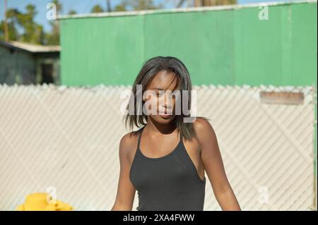 Una giovane donna con i capelli scuri che indossa una canotta nera si staglia all'aperto, stringendo leggermente sotto il sole, con un edificio verde e una recinzione a catena sullo sfondo. Foto Stock