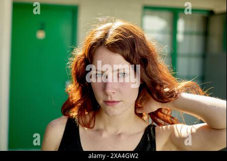 Una donna dai capelli rossi con occhi verdi sorprendenti è pronta davanti a una porta verde, la sua mano che corre tra i capelli. Foto Stock