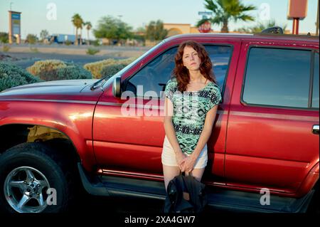 Una donna si appoggia su un pick-up rosso in una giornata di sole con un cielo azzurro e un verde paesaggistico sullo sfondo. Foto Stock