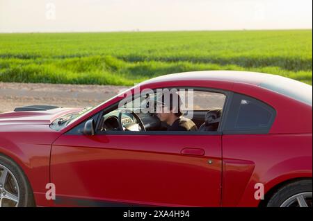 Una persona che indossa occhiali da sole e un berretto sta guidando un'auto sportiva rossa attraverso un'area di campagna con campi verdi sullo sfondo durante il giorno. Foto Stock