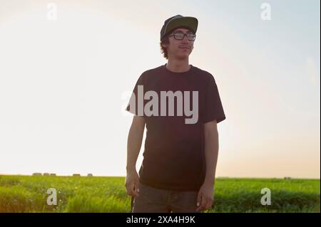 Un giovane si trova in un campo soleggiato con una t-shirt nera, occhiali da sole e un berretto, con un leggero sorriso sul viso. Foto Stock