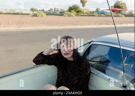 Una donna siede nella finestra aperta di un'auto d'epoca, appoggiando il gomito sul tetto, con un paesaggio desertico sullo sfondo. Foto Stock