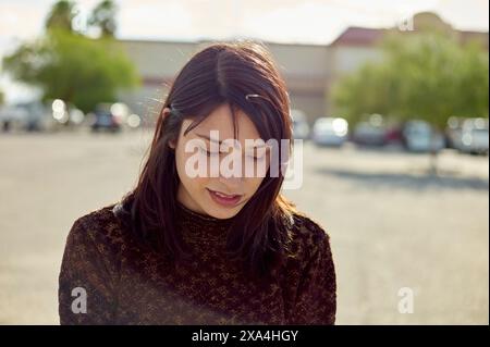 Una donna con i capelli scuri sta all'aperto in una giornata di sole, il suo sguardo è rivolto verso il basso mentre la luce del sole filtra i capelli. Foto Stock