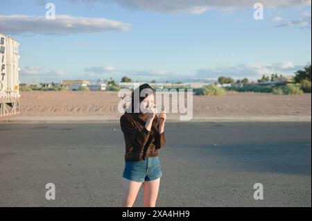 Una donna sta su un terreno sterrato, scattando una foto con il suo smartphone, con un cielo limpido e una vegetazione sparsa sullo sfondo. Foto Stock
