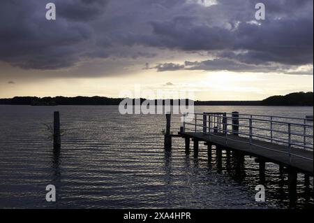 Un tranquillo molo si estende in un lago calmo sotto un cielo nuvoloso al crepuscolo, con il sole che sbircia attraverso le nuvole vicino all'orizzonte. Foto Stock