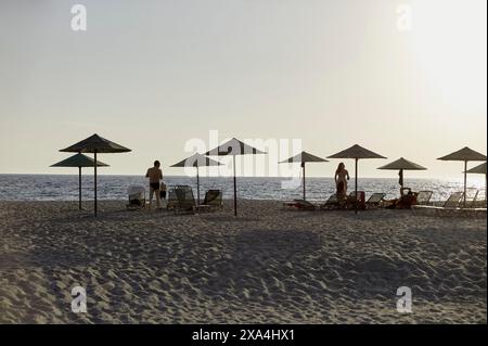 L'immagine raffigura una tranquilla scena di spiaggia al tramonto con diversi ombrelloni, sedie a sdraio e alcune persone che si godono l'atmosfera tranquilla. Foto Stock