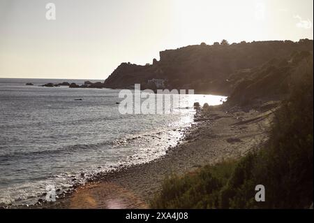 La luce del sole dorata bagna una spiaggia tranquilla con un dolce surf, incorniciata da una costa frastagliata e da un cielo limpido al crepuscolo. Foto Stock