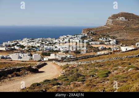 Una vista panoramica di un villaggio costiero con edifici bianchi sullo sfondo di cieli azzurri, con un sentiero sterrato che conduce attraverso un paesaggio collinare. Foto Stock