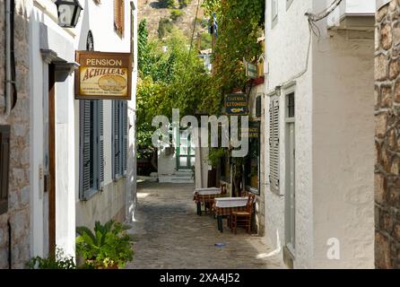 Un pittoresco vicolo acciottolato fiancheggiato da tradizionali edifici bianchi, con cartelli per una pensione e una taverna, e posti a sedere all'aperto sotto un baldacchino di foglie verdi. Foto Stock
