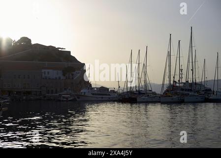 Tramonta su un porto calmo con sagome di barche a vela ancorate e yacht contro una collina con edifici, sotto un cielo che svanisce dolcemente. Foto Stock