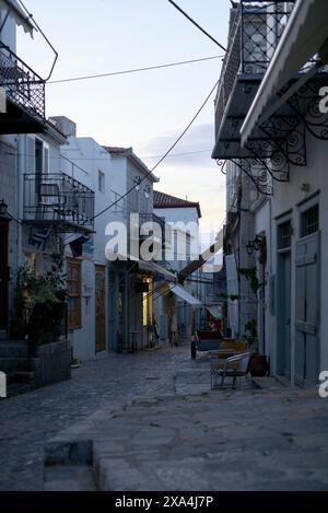 Una tranquilla strada acciottolata al tramonto, fiancheggiata da edifici tradizionali e balconi, con sedie e tavoli che suggeriscono un quartiere tranquillo. Foto Stock