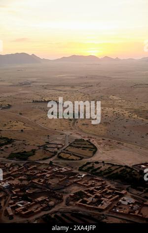 Una vista aerea di un insediamento con edifici in terra circondata da un vasto e arido paesaggio al tramonto, con montagne in lontananza e il sole che si tuffa sotto l'orizzonte. Foto Stock