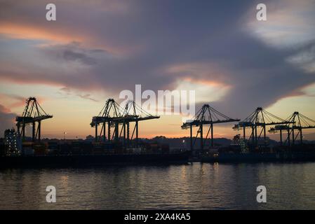 L'immagine mostra un porto al tramonto con profili sagomati di grandi gru container contro un cielo vibrante con sfumature arancioni e blu. Foto Stock