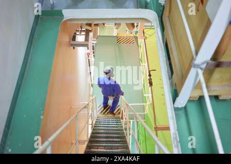 Una persona con tute blu sta scendendo una scala in metallo in un ambiente industriale con corrimano di sicurezza gialle. Foto Stock