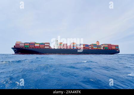 Una grande nave da carico è carica di contenitori colorati e naviga attraverso l'oceano blu sotto un cielo limpido. Foto Stock
