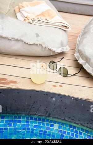 Una bevanda rinfrescante con una fetta di lime siede su una superficie di legno accanto ad una piscina, accompagnata da un paio di occhiali da sole e comodi cuscini. Foto Stock