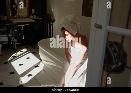 Una donna siede pensiamente accanto a una vasca indipendente in un bagno illuminato dal sole avvolto in un asciugamano con i capelli in un turbante. Foto Stock