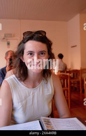 Una donna è seduta a un tavolo in un caffè ben illuminato, sorridendo leggermente alla macchina fotografica con un menu davanti a lei. Foto Stock