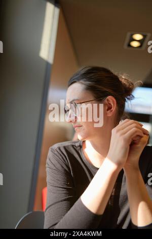 Una donna premurosa che indossa gli occhiali è seduta in casa con il mento appoggiato sulla mano, guardando lateralmente, illuminata dalla luce naturale. Foto Stock
