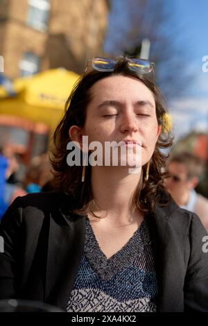 Una giovane donna con gli occhi chiusi gode della luce del sole sul viso, indossa un blazer nero su una parte superiore blu e gli occhiali da sole premuti sulla testa. Foto Stock