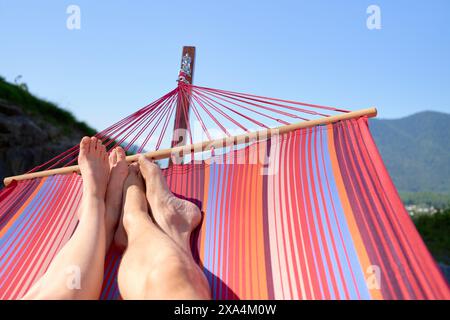 Due persone si rilassano in un'amaca a strisce rosse e arancioni all'aperto, con un cielo azzurro e una lussureggiante montagna verde sullo sfondo. Foto Stock