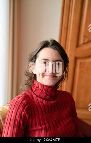 Una donna sorridente in un maglione dolcevita rosso è seduta all'interno accanto a una porta di legno, con luce naturale che le illumina il viso. Foto Stock