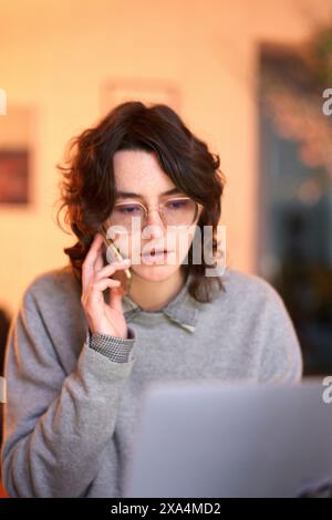 Un individuo che indossa gli occhiali lavora intensamente su un laptop mentre tiene una conversazione telefonica in un ambiente interno molto illuminato. Foto Stock