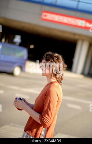 Una persona con i capelli ricci sorride e si trova in una zona illuminata dal sole, possibilmente in un parcheggio, con un cielo azzurro limpido sopra e un edificio con testo rosso sullo sfondo. Foto Stock