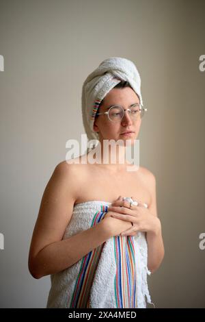Una giovane donna avvolta in un asciugamano in piedi su uno sfondo neutro, indossando occhiali, con un altro asciugamano avvolto intorno ai capelli. Foto Stock