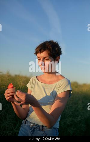 Una donna sta in piedi in un campo a ispezionare un piccolo oggetto rosso tra le sue mani durante l'ora d'oro, con la luce del sole che proietta una luce soffusa sulla scena. Foto Stock