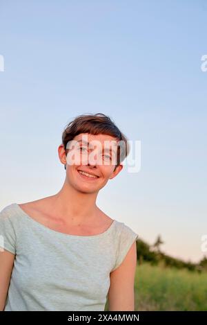 Una donna sorridente con i capelli corti, che indossa una parte superiore di colore chiaro, si erge all'aperto con un cielo limpido sullo sfondo. Foto Stock