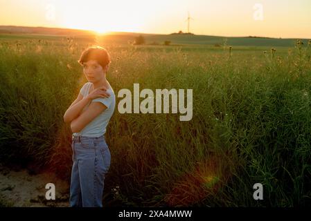 Una giovane donna si trova in un campo verde al tramonto, con il sole che proietta un caldo bagliore e crea uno sfondo sereno con turbine eoliche in lontananza. Foto Stock