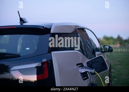 Un'auto elettrica si sta ricaricando in una stazione durante il crepuscolo, con il cavo di ricarica collegato alla porta sul lato del veicolo. Foto Stock